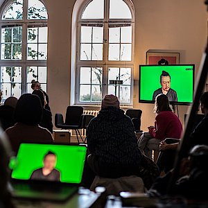 [Translate to Englisch:] Es sind zwei Bildschirme zu sehen. Der Laptop und der Fernseher, der den Inhalt des Laptops wiedergibt. Eine Menschengruppe schaut auf den Fernseher. Zu sehen ist ein Mensch auf grünem Hintergrund. Das Bild wurde während der Keynote von Choy Ka Fai gemacht.