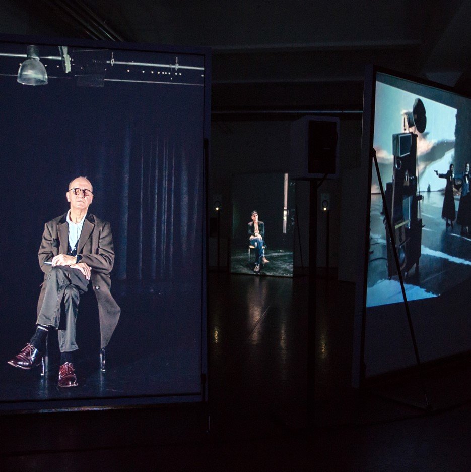 Auf mehreren Videotafeln in einer Ausstellung sind sitzende Menschen zu sehen. Der sitzende Mann im Vordergrund trägt einen langen Mantel und eine Schildpattbrille.